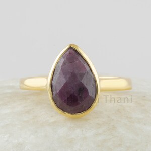 Pode incluir: Um anel de ouro com uma gema facetada de cor vermelha escura em forma de lágrima.