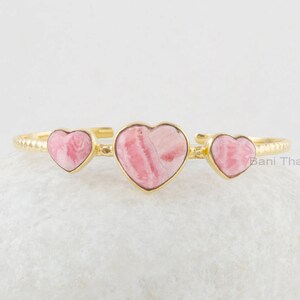 May include: A gold bracelet with three heart-shaped pink gemstones. The bracelet has a textured band. The gemstones have a unique pattern. The bracelet is displayed on a white, textured surface.