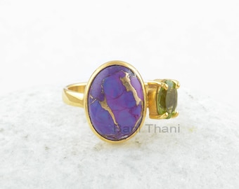 Pode incluir: Anel de ouro com uma gema oval de cor púrpura e dourada e uma gema oval verde menor. O anel está sobre uma superfície branca.