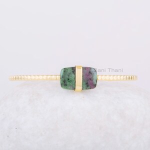 May include: A gold ring featuring a green and pink gemstone set in a gold band. The ring is displayed on a white rock.