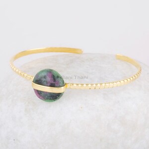 May include: Gold bangle bracelet featuring a green and pink gemstone. The bracelet has a textured design and the gemstone is set in a gold bezel.