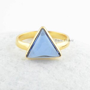 Pode incluir: Um anel de ouro com uma gema triangular azul claro. O anel está sobre um fundo branco.