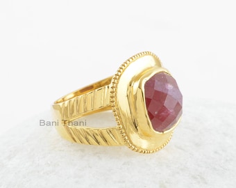 Könnte beinhalten: Goldring mit einem roten Edelstein, der in einer quadratischen Fassung sitzt. Der Ring hat eine strukturierte Band.