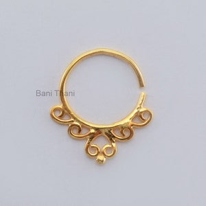 Puede incluir: Anillo de nariz de oro con un diseño decorativo y adornado. El anillo está abierto y tiene una pequeña perla redonda en la parte inferior.