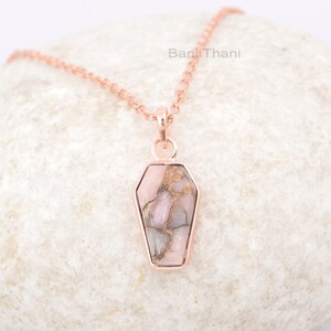 K&ouml;nnte beinhalten: Eine Halskette aus ros&eacute;goldener Kette mit einem Sarg-f&ouml;rmigen Anh&auml;nger, der mit einem rosa und braunen Opal-Stein verziert ist.