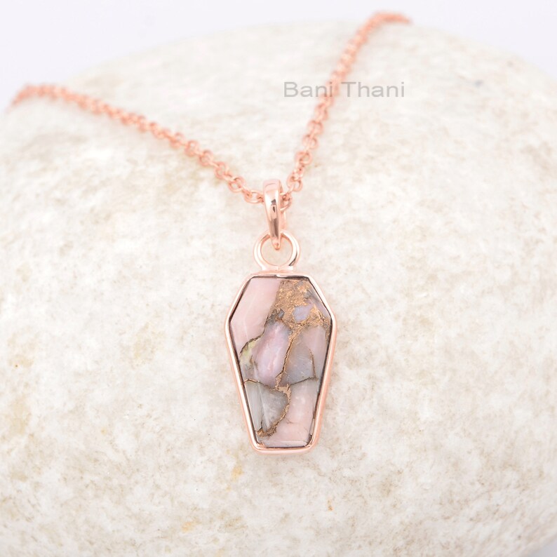 K&ouml;nnte beinhalten: Eine Halskette aus ros&eacute;goldener Kette mit einem Sarg-f&ouml;rmigen Anh&auml;nger, der mit einem rosa und braunen Opal-Stein verziert ist.