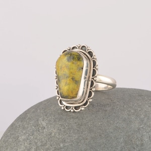 May include: A silver ring with a large, yellow and green gemstone set in a decorative silver bezel. The ring is on a gray rock.