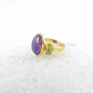 May include: Gold ring with a purple and gold oval stone and a smaller green oval stone. The ring is on a white surface.