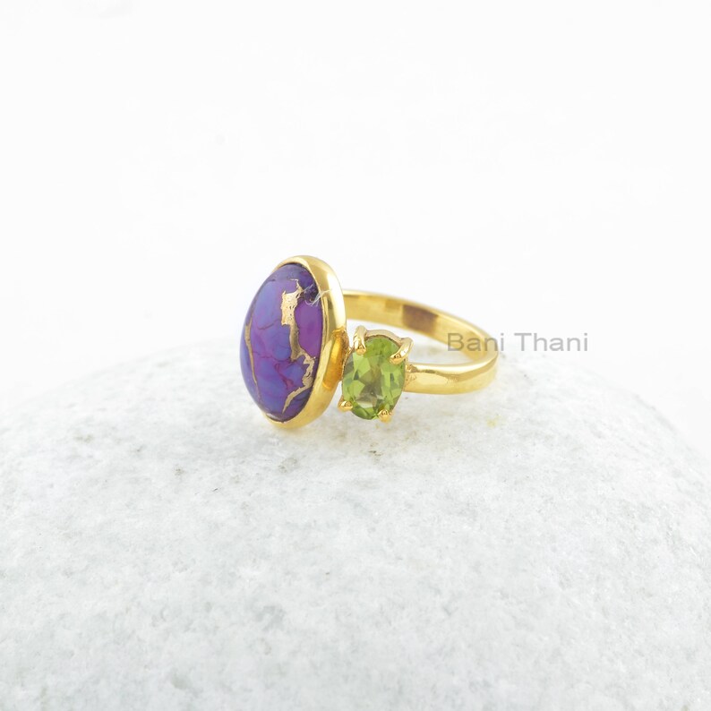 May include: Gold ring with a purple and gold oval stone and a smaller green oval stone. The ring is on a white surface.