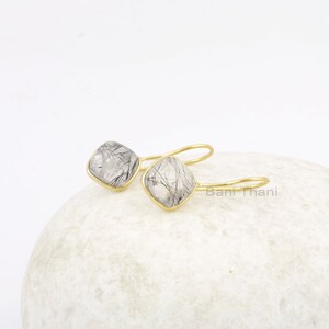 May include: A pair of gold-colored earrings with square, clear stones featuring black inclusions. The stones are set in a gold bezel and attached to gold-colored ear wires. The earrings are displayed on a white, textured surface.