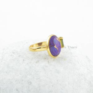 May include: A gold ring with a purple and gold oval gemstone and a smaller green gemstone. The ring is on a white surface.
