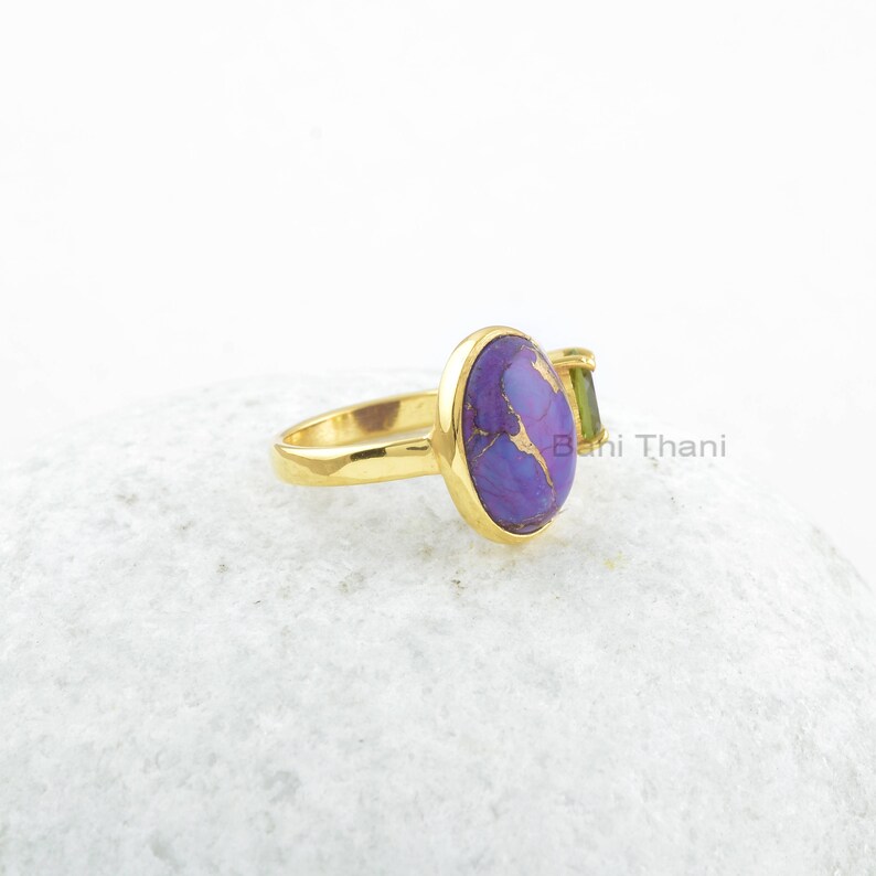 May include: A gold ring with a purple and gold oval gemstone and a smaller green gemstone. The ring is on a white surface.