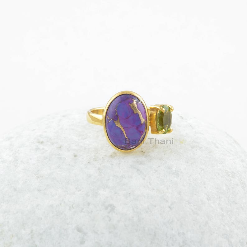May include: Gold ring with a purple and gold oval gemstone and a smaller green oval gemstone. The ring is on a white surface.