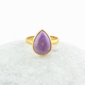 Op de afbeelding: Een gouden ring met een paarse, traanvormige edelsteen. De ring ligt op een witte achtergrond.