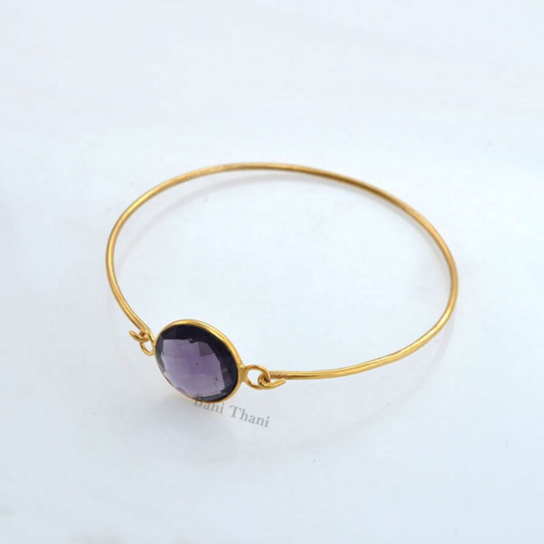 May include: A gold bangle bracelet with a single round, dark purple gemstone. The bracelet is open and has a simple design.
