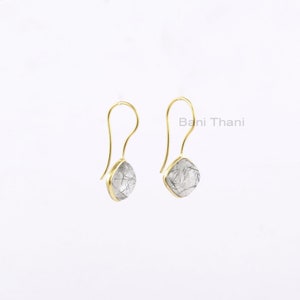 May include: A pair of gold-colored drop earrings. Each earring features a teardrop-shaped, clear stone with black inclusions, set in a gold-colored bezel. The earrings have a simple, curved hook design. The text "Bani Thani" is visible.
