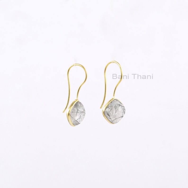 May include: A pair of gold-colored drop earrings. Each earring features a teardrop-shaped, clear stone with black inclusions, set in a gold-colored bezel. The earrings have a simple, curved hook design. The text "Bani Thani" is visible.