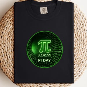 May include: Black t-shirt with a green graphic of the Pi symbol, the numbers 3.14159, and the words "PI DAY". The design is inside a green circle with a binary code background. The shirt is displayed on a woven, circular mat.