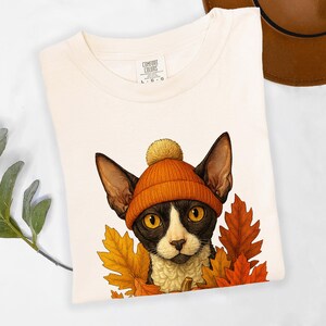 May include: Cream-colored t-shirt featuring a cat wearing an orange beanie and surrounded by autumn leaves. The cat has black and white markings and large yellow eyes. The shirt has a Comfort Colors tag.