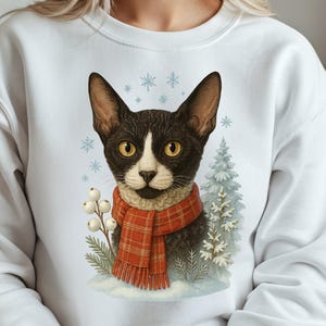 May include: White sweatshirt featuring a black and white cat wearing a red plaid scarf. The cat is surrounded by winter elements like snowflakes and a small evergreen tree. The design has a watercolor style.