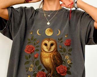 May include: Dark gray t-shirt featuring a detailed illustration of a barn owl surrounded by red roses, crescent moons, and a full moon. The owl has brown and tan feathers. The design includes small yellow accents and floral elements.