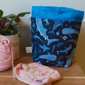 May include: A blue and black whale shark print drawstring project bag with a blue top. A ball of pink yarn with knitting needles and a partially knitted sock are next to the bag. A small plant in a white pot is in the background.