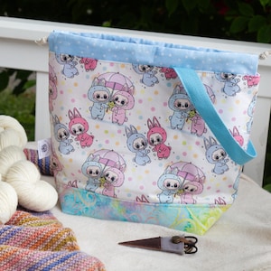 May include: A drawstring project bag featuring a white fabric with a repeating pattern of cartoon characters in pink and blue bunny suits holding umbrellas, accented by a light blue polka dot top band and a colorful marbled bottom panel.
