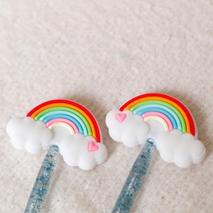 May include: Two rainbow-colored silicone phone grips with white clouds and pink hearts. The grips are attached to clear glitter phone cases.