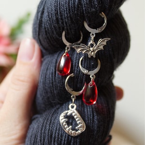 May include: Silver-toned stitch markers with a vampire theme. Each marker features a different charm: a bat, a set of fangs, and red teardrop-shaped beads. The markers are attached to a dark blue skein of yarn.