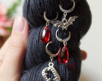 May include: Silver stitch markers with a vampire theme. Each marker has a different charm: a bat, fangs, and red teardrop beads. The markers are attached to a dark blue skein of yarn.