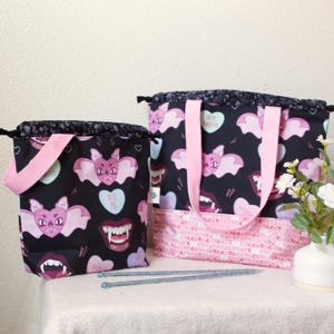 May include: Two black tote bags with pink accents. The bags feature a repeating pattern of pink bats, vampire teeth, and heart-shaped candies with the text "Bite Me" on them. One bag has a drawstring closure, and the other has pink handles.