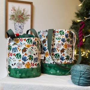 May include: Two white fabric project bags with a colorful sweater pattern. The bags have green handles and a green base with a snowflake design. One bag is open, revealing a green interior. A ball of teal yarn sits nearby.