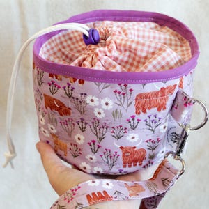 May include: A cylindrical fabric project bag featuring a light purple exterior with a pattern of orange Highland cows, white flowers, and pink thistles. It has a white drawstring closure with a purple toggle and a peach gingham fabric lining.