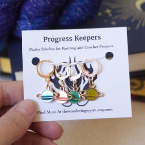 May include: A card with four planet-shaped progress keepers for knitting and crochet projects. The keepers are gold-toned with colorful enamel planets in teal, coral, green, and yellow. The card reads "Progress Keepers" and "Marks Stitches for Knitting and Crochet Projects."
