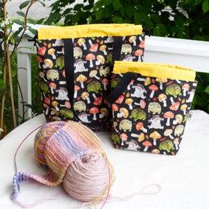 May include: Two black fabric bags with a whimsical pattern of frogs and mushrooms. The bags have yellow lining and black handles. A ball of yarn and knitting needles are in the foreground.