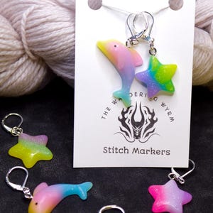 May include: A set of colorful stitch markers featuring a dolphin and a star design. The markers are made of resin and have a gradient of pink, yellow, blue, and green. The markers are attached to silver rings. The card reads "The Wandering Wyrm Stitch Markers".