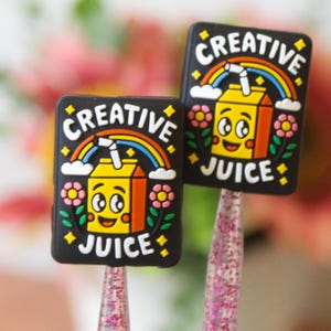 May include: Two black rectangular charms with the words "CREATIVE JUICE" and a cartoon juice box with a rainbow, clouds, and flowers. The charms are on pink glittery sticks.
