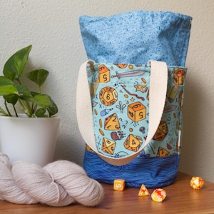 May include: A light blue tote bag with a cream-colored handle, featuring a pattern of orange dice, swords, and stars. The bag's interior is a lighter blue with a swirl design. Several dice are scattered on the wooden surface.