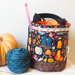 May include: A black fabric yarn tote bag with a colorful autumn-themed pattern featuring pumpkins, boots, and gardening tools. A pink crochet hook and measuring tape are visible. A ball of blue yarn and a pumpkin are next to the bag.