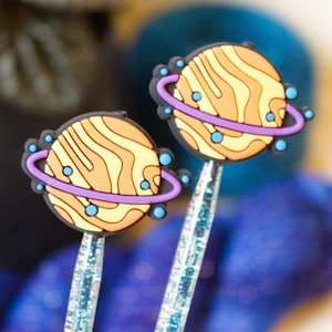 May include: Two sparkly blue pens with orange and yellow planet toppers. The planets have purple rings and blue dots.