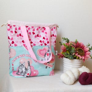 May include: A pink and blue tote bag with a cat graphic and the text "Razzle Dazzle". The bag has pink handles and a pink heart pattern. The bag is made of fabric and has a drawstring closure.