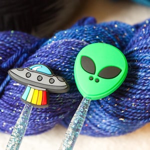 May include: Two knitting needles with colorful glitter shafts and plastic toppers. One topper is a green alien face and the other is a gray UFO with a rainbow stripe.