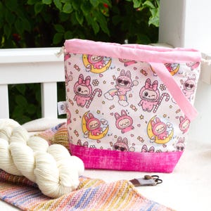 May include: A drawstring project bag featuring a white fabric with a repeating pattern of pink cartoon bunnies, yellow crescent moons, rainbows, and sweets. The bag has a solid pink fabric base, a pink drawstring top closure, and a pink fabric handle.