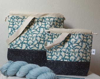 May include: Two drawstring project bags featuring a teal and cream cat pattern with a solid black base. The bags have cream-colored fabric handles. A skein of light blue yarn and two knitting needles are positioned in front of the bags.