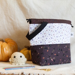 May include: A fabric drawstring project bag featuring a two-tone design with a white upper section patterned with small pumpkins and a dark brown lower section patterned with witch hats and cauldrons, complete with a black handle and drawstring.