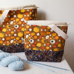May include: Two fabric project bags with a pumpkin pattern. The bags have a brown background with orange and yellow pumpkins. The bottom of the bags have a floral pattern. One bag has a handle. Light blue yarn and knitting needles are in the foreground.