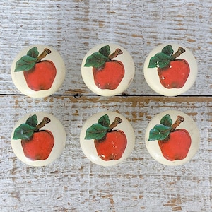 Op de afbeelding: Zes witte keramische knoppen met rode appelmotieven. De appels hebben groene bladeren en stelen.