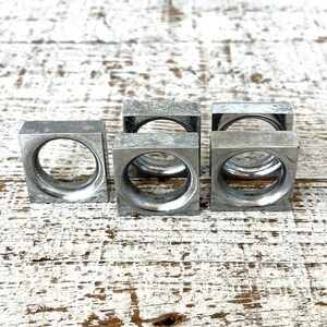 May include: Five silver metal napkin rings in a square shape with a round opening in the centre. The rings are arranged in a row on a white wooden surface.