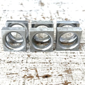 Op de afbeelding: Vijf zilveren metalen servetringen, elk met een vierkante vorm en een ronde opening in het midden. De ringen zijn in een rij op een wit houten oppervlak gerangschikt.