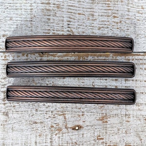 Puede incluir: Cuatro tiradores de armario de bronce antiguo con diseño de cuerda. Los tiradores están dispuestos en fila sobre una superficie de madera blanqueada.